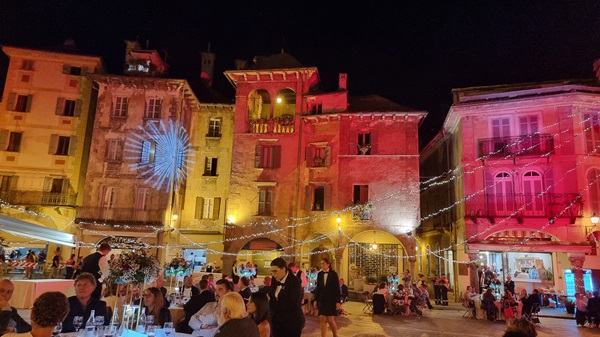 Piazza Mercato in centro a Domodossola illuminata da giochi di luce, con tavoli apparecchiati per la cena di beneficenza Domosofia Gourmet 2024